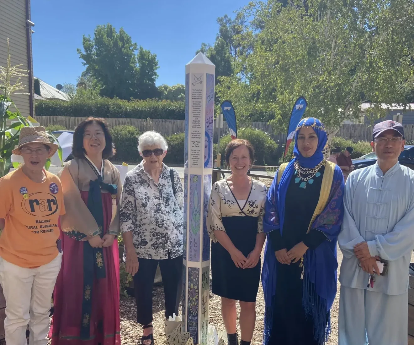 Diverse group standing around peace pole outdoors.