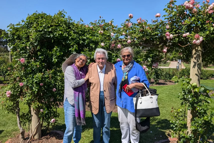 Three adults smiling in garden under rose arch.