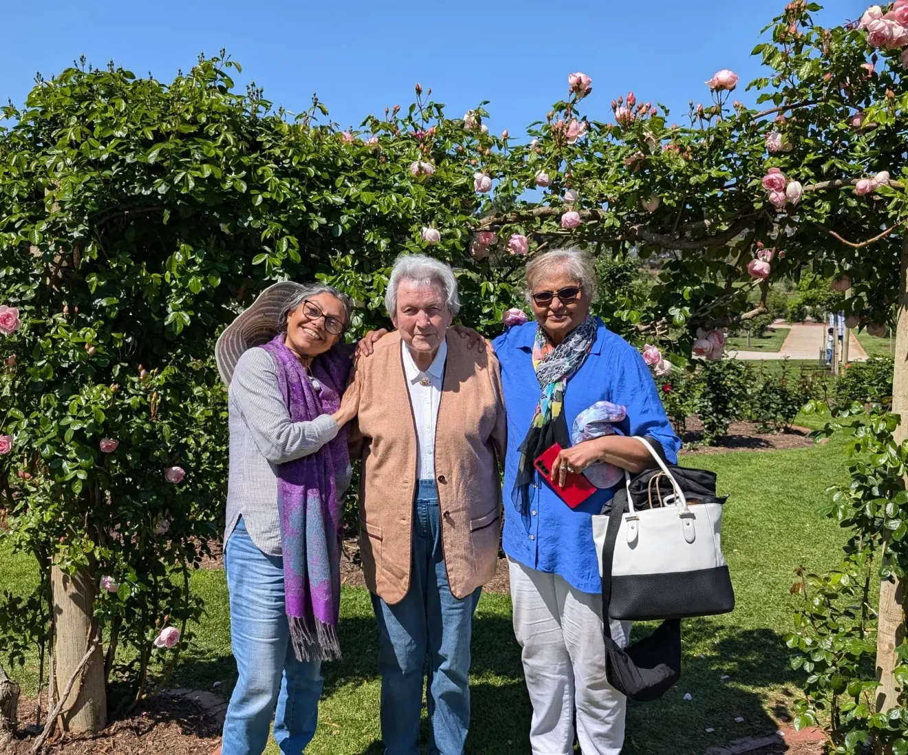 Three adults smiling in garden under rose arch.